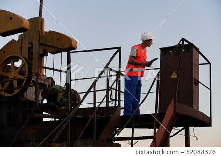 Male worker in protective helmet holding clipboard while monitoring work of petroleum pump jack. Petroleum engineer in work vest working in oil field. Concept of petroleum industry and oil extraction. 82088326