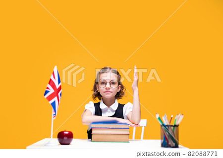 Serious caucasian schoolgirl sitting at the desk and raising hand. English lesson Great Britain flag Serious caucasian schoolgirl sitting at the desk and raising hand. English lesson Great Britain flag 82088771
