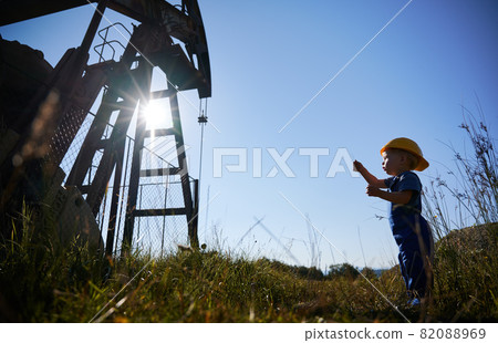 Side low view of child boy who standing opposite metal fence and examining oil pump structure in summer sunny day. On the background cloudless blue sky. Side low view of child boy who standing opposite metal fence and examining oil pump structure in summer sunny day. On the background cloudless blue sky. 82088969