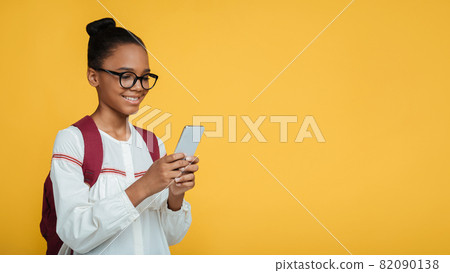 Cheerful smart young afro american lady pupils in glasses with backpack typing message on smartphone Cheerful smart young afro american lady pupils in glasses with backpack typing message on smartphone 82090138