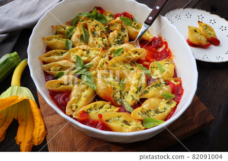 Big shell shape pasta stuffed with creamy soft cheese, zucchini and spinach sprinkled with parmesan cheese in baking dish wooden table, healthy dinner recipe, view flat lay . 82091004