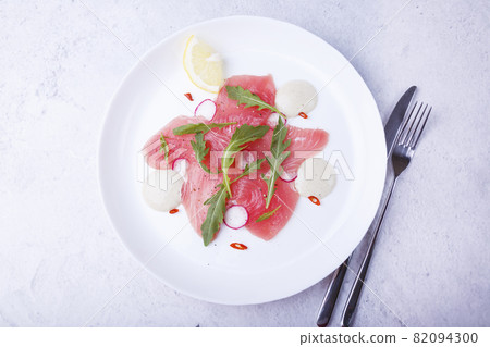 Fresh tuna carpaccio with arugula, radish, chili, lemon and sauce. Traditional cold appetizer of Italian cuisine. Close-up, selective focus, top view. 82094300