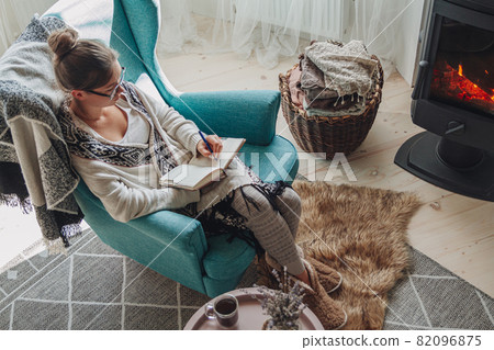 Young woman write in notebook sitting in armchair by fireplace Young woman write in notebook sitting in armchair by fireplace 82096875