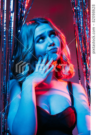Bad party. Thoughtful woman. New year celebration. Holiday fail. Bored disappointed lady looking up hand on cheek in shimmering silver cascade curtain blue red neon light. Bad party. Thoughtful woman. New year celebration. Holiday fail. Bored disappointed lady looking up hand on cheek in shimmering silver cascade curtain blue red neon light. 82098628