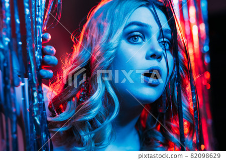 Curious woman. Christmas party. Festive surprise. Holiday expectation. Pretty interested lady curly hair looking from shimmering silver cascade curtain blue red neon light. 82098629