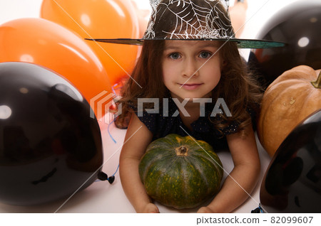 Close-up portrait of pretty little girl in carnival witch costume and wizard hat looking at camera posing with pumpkin in the white background with colored black and orange balloons. Halloween concept 82099607