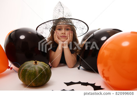 Pretty little witch girl in a wizard hat lying on a white background with copy space next to handmade felt bats, pumpkin and orange black balloons. Traditional event, Halloween party concept. 82099608
