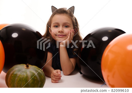Traditional event, Halloween party concept. Beautiful little witch girl in hoop with cat ears lying on white background with copy space next to pumpkin and orange black balloons Traditional event, Halloween party concept. Beautiful little witch girl in hoop with cat ears lying on white background with copy space next to pumpkin and orange black balloons 82099610