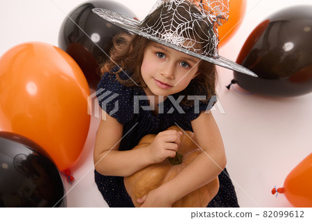 4 years old adorable pretty little girl in witch dress and wizard hat looks at camera playing with balloons and a pumpkin isolated on white background. Concept of a child having fun at Halloween party 82099612