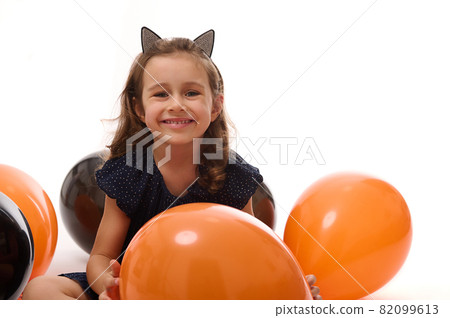 Portrait of cute witch girl in Halloween dress and hoop with cat ears having fun with colorful black and orange balloons isolated on white background with copy space. Halloween party. 82099613