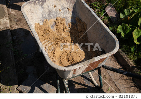 A garden wheelbarrow filled with sand and dry cement mixture stands on the path A garden wheelbarrow filled with sand and dry cement mixture stands on the path 82101873