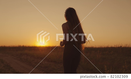 A girl in headphones runs along the road at sunset, a young woman makes a morning jog, a healthy lifestyle outdoors, a long-distance race, preparation for victory in a sports competition 82102309