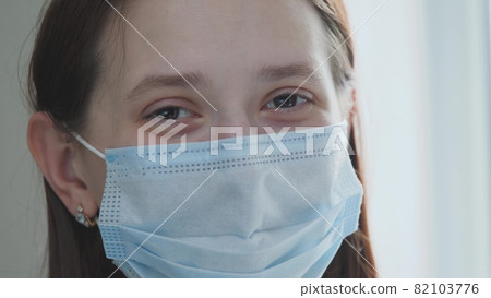 Young girl puts on a medical mask while standing by the window, the female is breathing deeply and smiling, looking at the camera, healthcare and medical concept, close-up portrait, 4k Young girl puts on a medical mask while standing by the window, the female is breathing deeply and smiling, looking at the camera, healthcare and medical concept, close-up portrait, 4k 82103776