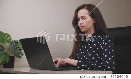 Serious business woman is sitting at the computer and typing a document on the keyboard, looking at the camera, the freelancer girl is busy, the concept of a happy business project on the online 82104046