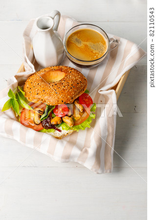 Breakfast with bagel, stuffed with salad leaves, mussels and tomatoes. With a cup of coffee and a creamer. On a wooden tray. Vertical 82110543
