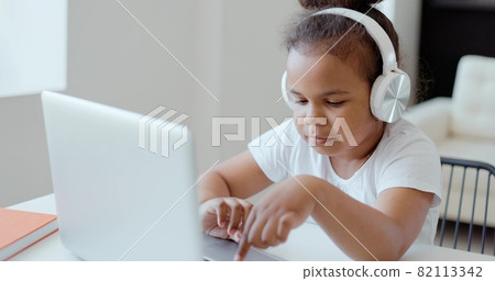 Afro american kid girl school pupil wearing headphones studying online from home watching web class lesson or listening tutor by video call elearning on pandemic isolation. Children remote education. Afro american kid girl school pupil wearing headphones studying online from home watching web class lesson or listening tutor by video call elearning on pandemic isolation. Children remote education. 82113342