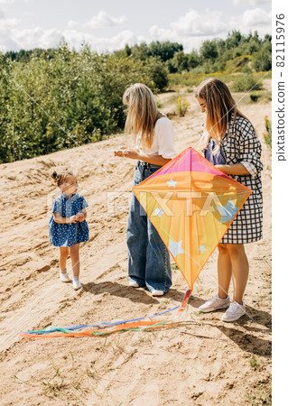A cute little girl together with her mother and sister are preparing a kite for launch during a vacation outside the city A cute little girl together with her mother and sister are preparing a kite for launch during a vacation outside the city 82115976