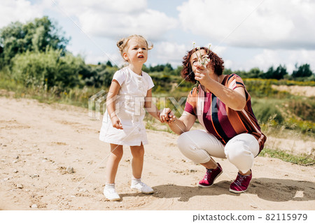 Grandmother and granddaughter are walking along a field road outside the city. An elderly woman is playing with a little girl Grandmother and granddaughter are walking along a field road outside the city. An elderly woman is playing with a little girl 82115979