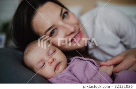 Front view of happy young mother with her newborn baby girl lying on sofa indoors at home, looking at camera. 82121026