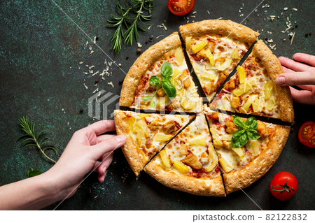 Hot Hawaiian pizza with chicken, pineapple and cheese, hands, taking pieces pizza served with cheese sauce. Delicious traditional Italian pizza on on a dark stone or concrete background. Top view flat Hot Hawaiian pizza with chicken, pineapple and cheese, hands, taking pieces pizza served with cheese sauce. Delicious traditional Italian pizza on on a dark stone or concrete background. Top view flat 82122832