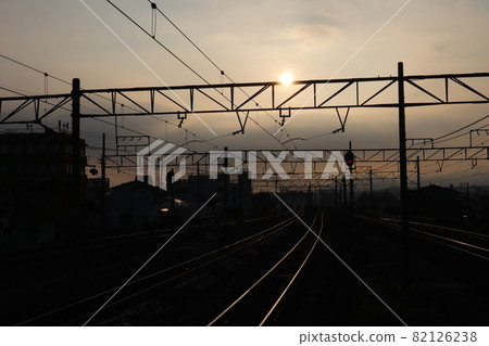 A landscape where the entire city with overhead lines, railroad tracks, buildings, and mountains looks dark due to the morning sun, gray clouds, the sky colored by the sunrise, and the backlight. 82126238
