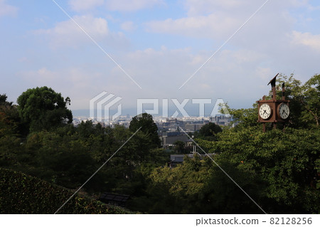 A forest of green trees, a clock, a city in Kyoto that can be seen in the distance, a mountain that can only be seen below with clouds, clouds of various shapes and colors, and a blue sky 82128256