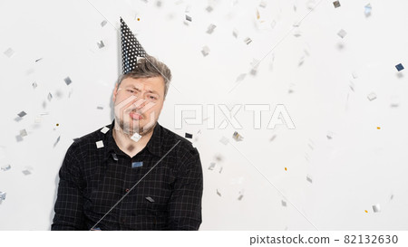 Bad party. Lonely Birthday. Ruined celebration. Introvert personality. Disappointed sad man in festive hat with confetti fall isolated on copy space white banner background. Bad party. Lonely Birthday. Ruined celebration. Introvert personality. Disappointed sad man in festive hat with confetti fall isolated on copy space white banner background. 82132630
