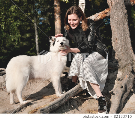 front view of young smiling woman stroking her dog whle sitting under the tree in a forest. walking with dog, pet friend. 82133710