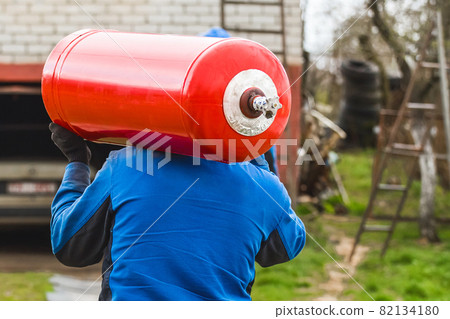 A male industrial worker walks with a gas cylinder to a gas car. Transportation and installation of a propane bottle to residential buildings 82134180