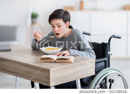 Focused disabled teenage boy in wheelchair eating cereal for breakfast and reading book at home. Domestic hobbies 82136016