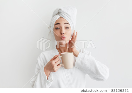 Smiling cute millennial caucasian blonde lady in bathrobe and towel on head, holds cup, drinks coffee 82136251