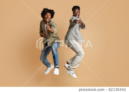 Two Cheerful Black Guys Pointing At Camera While Jumping Over Beige Background 82136342
