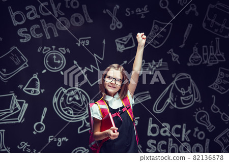Happy schoolgirl in glasses preparing to go to school holding book raising hand up. Back to school  82136878
