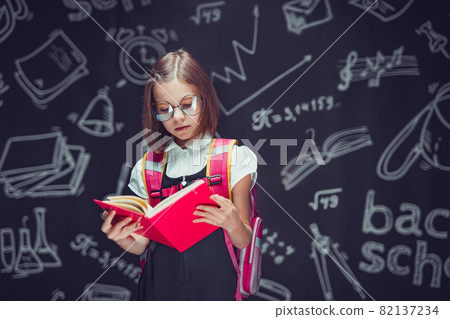 Serious schoolgirl in glasses preparing to go to school with backpack reading book. Back to school Serious schoolgirl in glasses preparing to go to school with backpack reading book. Back to school 82137234