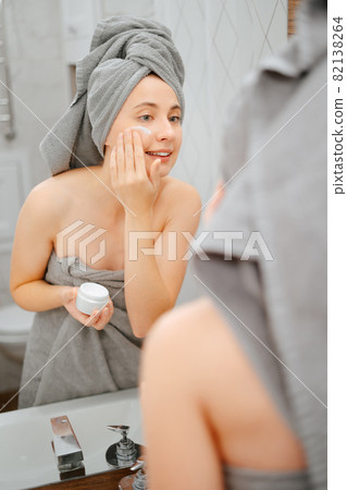 Woman with a towel on her head after a shower, apply a moisturizer on her face. Woman with a towel on her head after a shower, apply a moisturizer on her face. 82138264