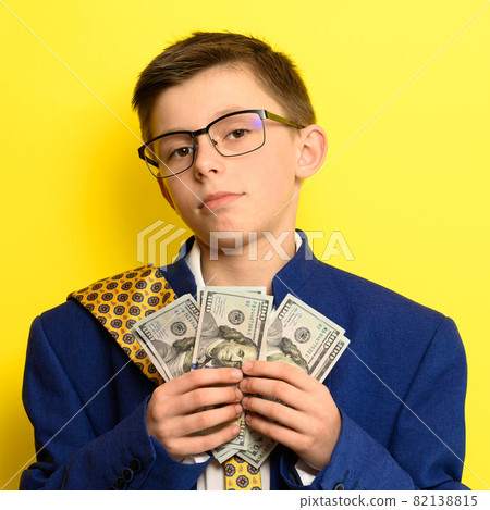 A boy in glasses and an oversized suit with a satisfied face is holding US dollars, a child and big money. A boy in glasses and an oversized suit with a satisfied face is holding US dollars, a child and big money. 82138815