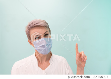 The medic points his finger at a place free for text or images. Doctor, blond woman with short hair. A doctor wearing a disposable medical mask The medic points his finger at a place free for text or images. Doctor, blond woman with short hair. A doctor wearing a disposable medical mask 82138905