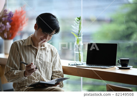 Smiling young man sitting bright office and making note on notebook. Smiling young man sitting bright office and making note on notebook. 82149508