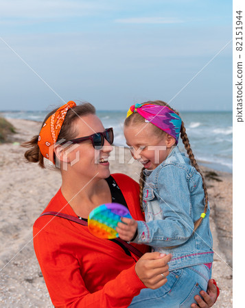 Cheerful mother and daughter smiling walking on sea beach. Happy family. Cute little child playing rainbow pop it toy 82151944