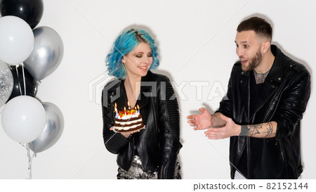 Birthday fun. Happy friends. Festive surprise. Party celebration. Funny shocked man wishing holiday cake with candle lights in hands of playful woman isolated white black silver balloons decor. Birthday fun. Happy friends. Festive surprise. Party celebration. Funny shocked man wishing holiday cake with candle lights in hands of playful woman isolated white black silver balloons decor. 82152144
