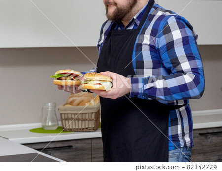 a man with a beard and in an apron holds burgers in his hands in the kitchen, background a man with a beard and in an apron holds burgers in his hands in the kitchen, background 82152729