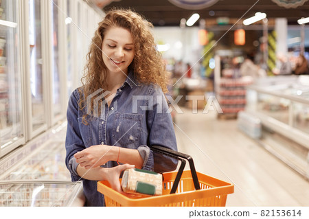 People, shopping and buying concept. Adorable beautiful female with curly hair, dressed in fashionable denim jacket, puts product in shopping backet, poses in supermarket with many products. 82153614