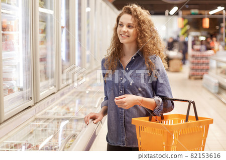 Cheerful adorable curly woman in denim jacket, holds shopping basket, strolls in supermarket, being in good mood. Pretty female comes at grocer`s shop to buy necessary products 82153616