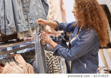 People, purchasing and shopping concept. Lovely curly woman wears comfortable denim jacket, holds hangers with jeans, going to buy new trousers, chooses suitable size and price. Female shopaholic 82153629