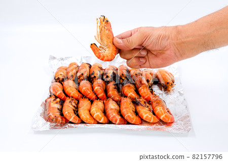 Close up a human hand picks up grilled river prawns from a pile on foil on white background. Close up a human hand picks up grilled river prawns from a pile on foil on white background. 82157796