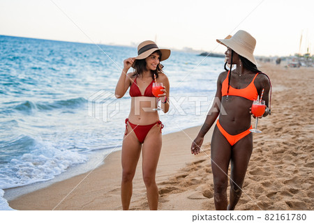 Young fit multiracial girlfriends in colorful swimwear and straw hats with glasses of refreshing beverages smiling and looking at each other while strolling together on sandy beach during summer Young fit multiracial girlfriends in colorful swimwear and straw hats with glasses of refreshing beverages smiling and looking at each other while strolling together on sandy beach during summer 82161780