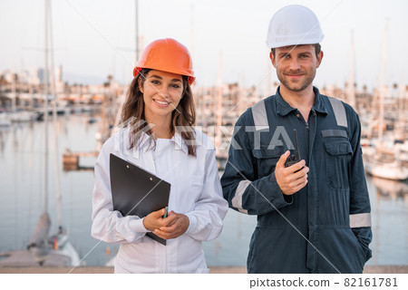 Cheerful young male and female logistic managers in hardhats and protective uniform with clipboard and VHF radio looking at camera while controlling shipment in sea port Cheerful young male and female logistic managers in hardhats and protective uniform with clipboard and VHF radio looking at camera while controlling shipment in sea port 82161781
