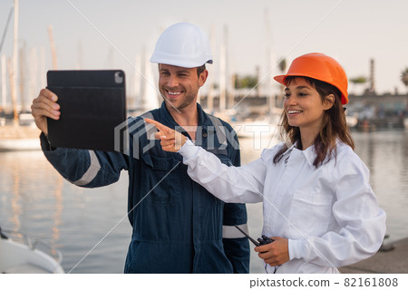 Optimistic young male and female engineers in helmets using tablet and discussing draft while inspecting sea port together 82161808