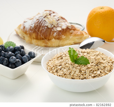 Raw oatmeal in white ceramic plate, blueberry, orange, on white table, Raw oatmeal in white ceramic plate, blueberry, orange, on white table, 82162892