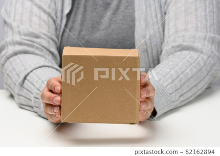 two female hands hold a square box made of brown corrugated cardboard on a white background, close up two female hands hold a square box made of brown corrugated cardboard on a white background, close up 82162894